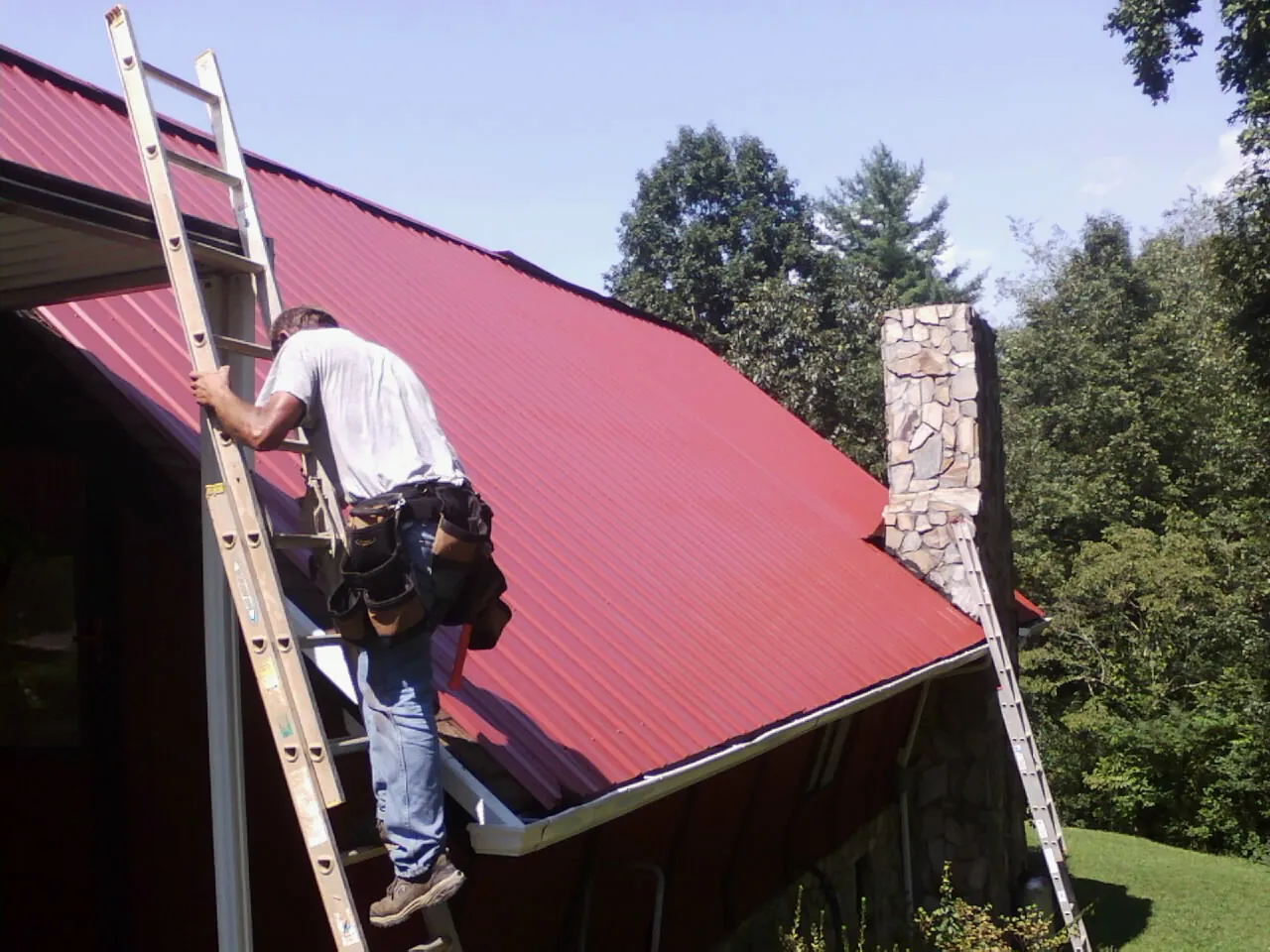Roofer on a ladder beside a red metal roof and stone chimney — Soffit & Fascia Repair by Roofing Brothers in Delta City